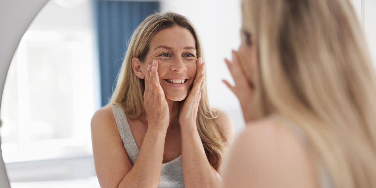 Kvinna tittar på sin spegelbild, rör vid sitt ansikte med sina händer och ler.