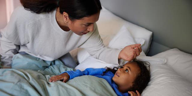 Mamma känner av temperaturen på sitt barn med handen mot barnets panna. Barnet ligger i sängen och kanske har feber och är sjuk.