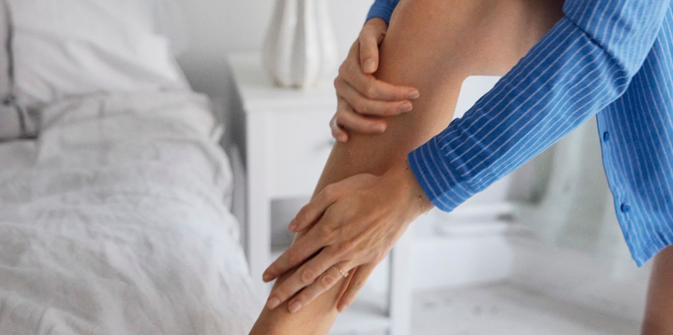 Person står brevid en en säng iförd en blå randig skjorta och applicerar hudkräm på sitt ben.