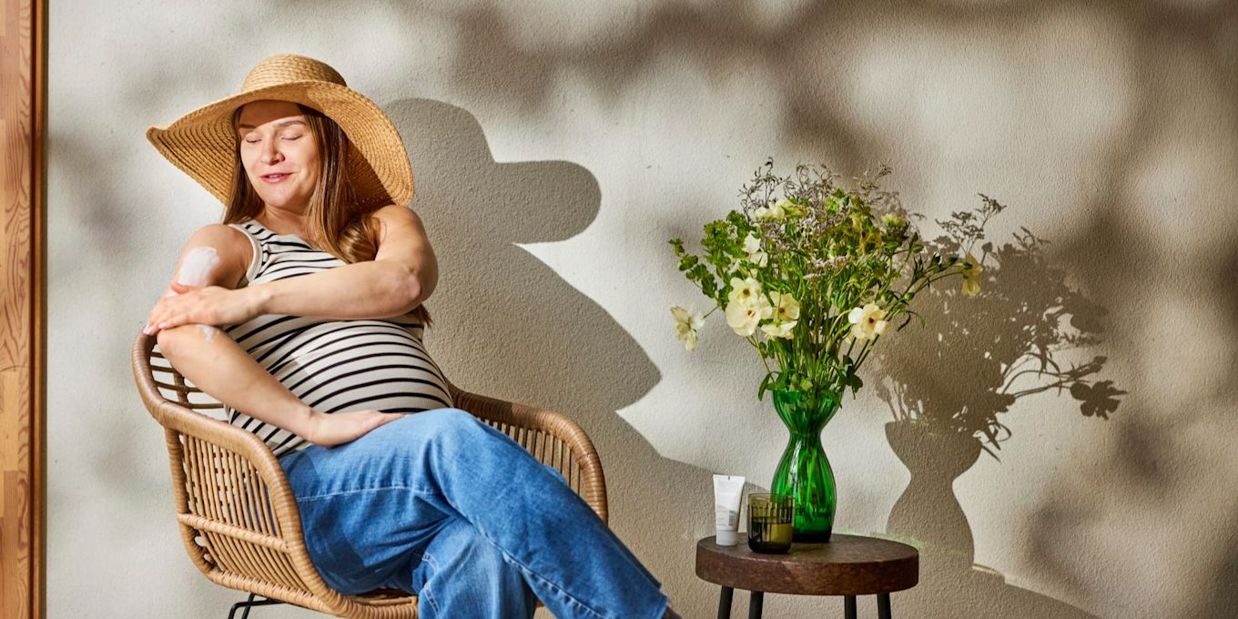 Gravid person sitter utomhus i solen och smörjer in armen med solskyddskräm. Bilden illustrerar hur du kan skydda huden vid solexponering under graviditeten.