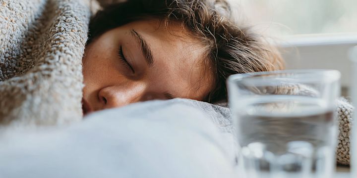 Sjuk tonåring, sovandes och nedbäddad i sängen med täcket långt uppdraget. Ett glas vatten syns på sängbordet. 