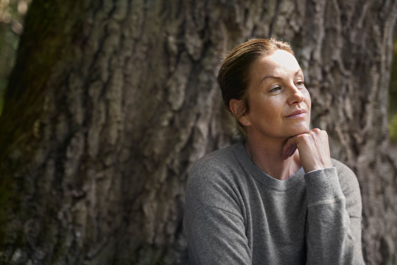 Kvinna sitter utomhus framför ett stort träd och tittar åt sidan.