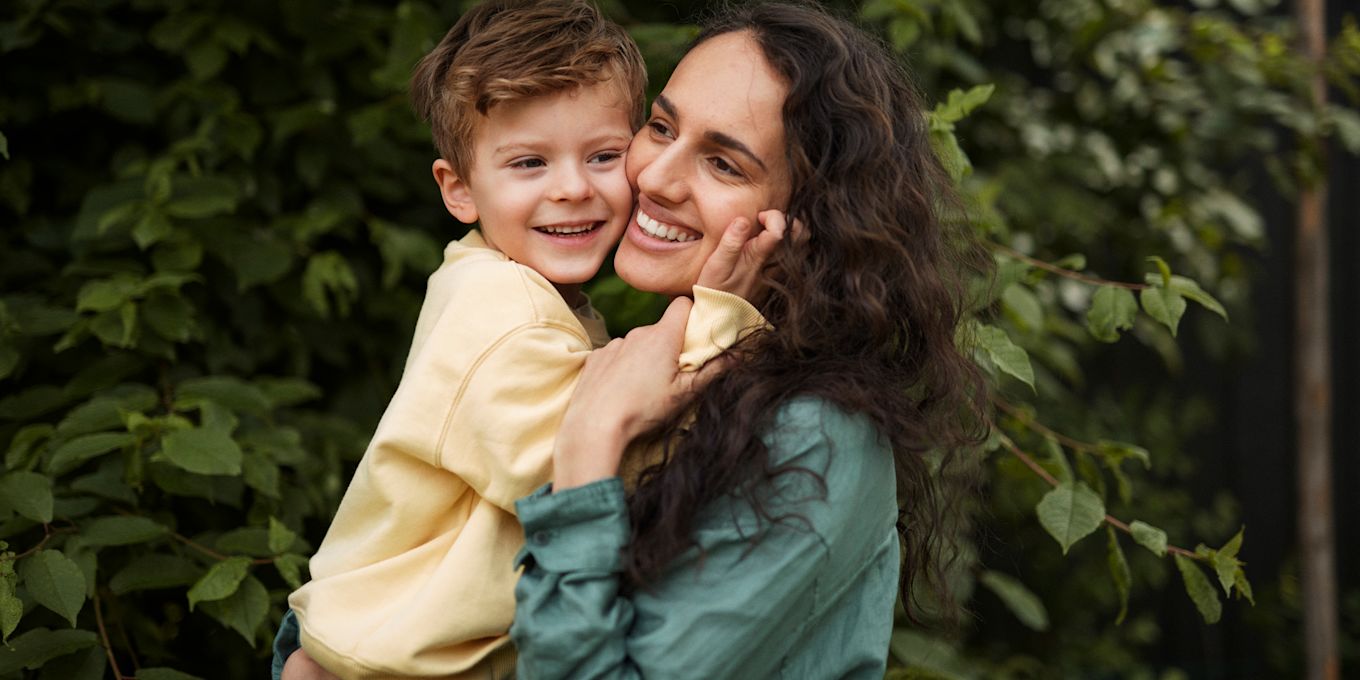 Mamma och son står utomhus framför en häck, mamma bär sonen i famnen och de kramas och ler.