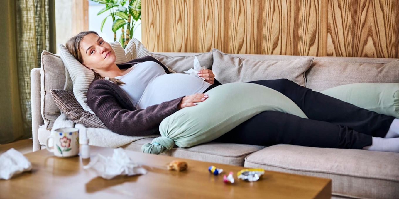 Gravid person vilar på en soffa med stöd av en gravidkudde och har näsdukar nära till hands. Bilden illustrerar egenvård och återhämtning vid förkylning under graviditeten.