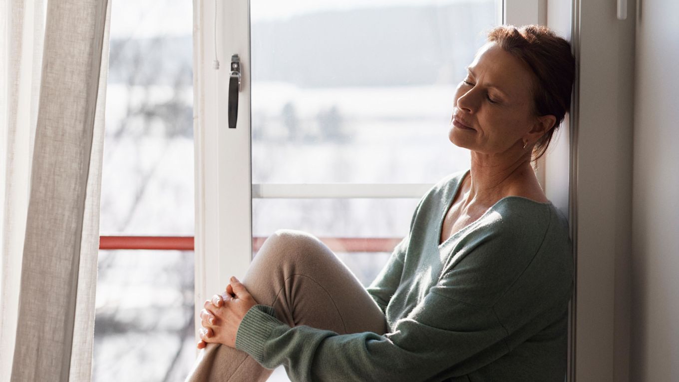 Kvinna sitter på en fönsterbräda och vilar med slutna ögon, solen lyser in genom fönstret.