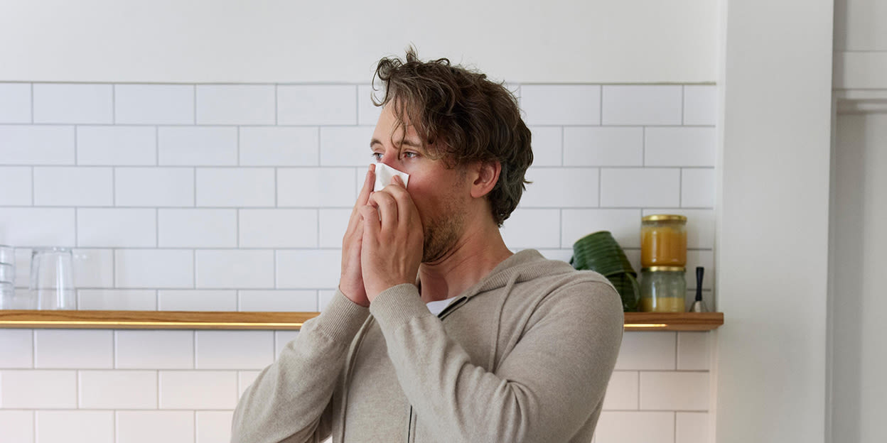 Förkyld kille står i köket och snyter sig med båda händerna.