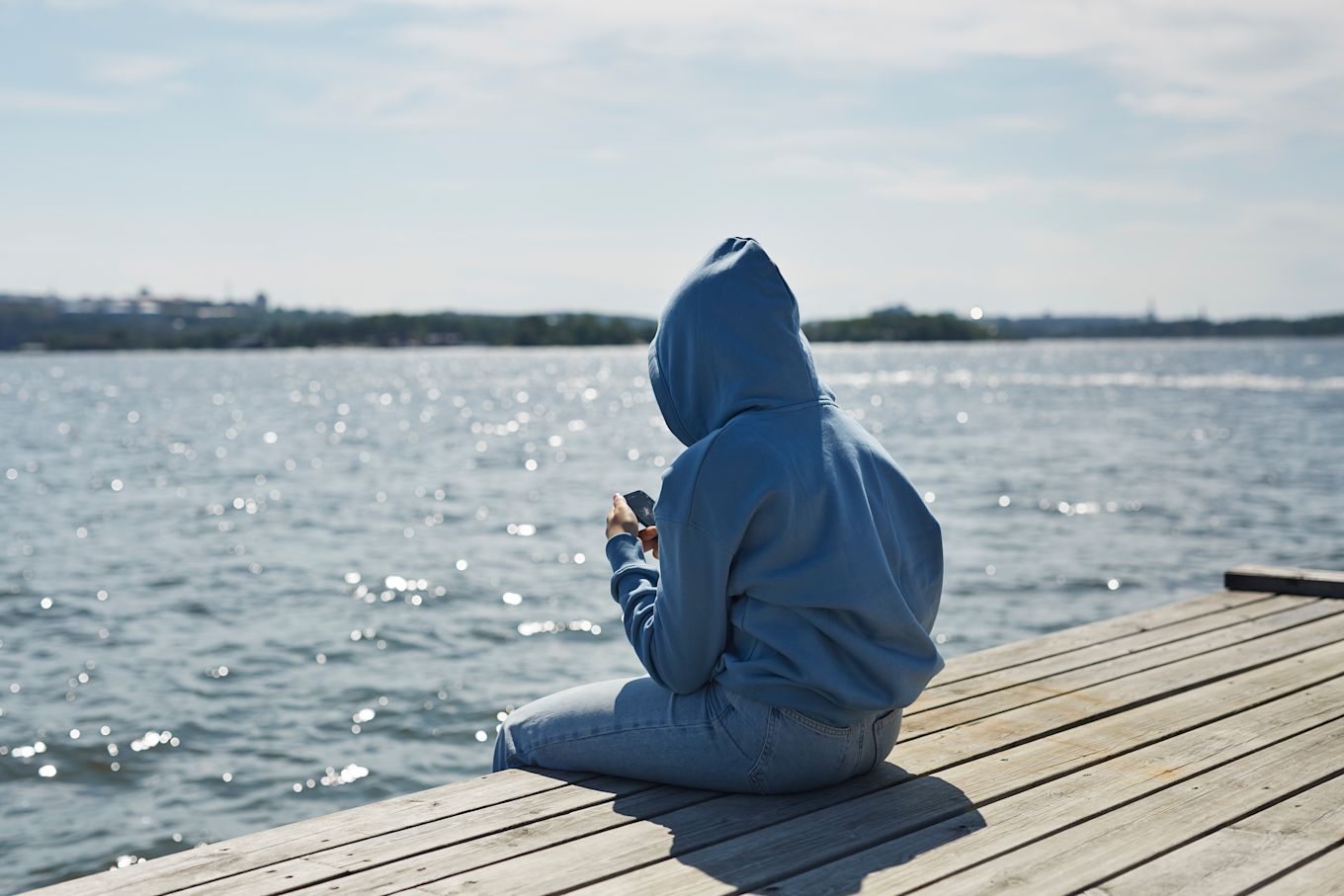 Tonåring sitter på en brygga i solskenet, med luvan uppdragen och tittar på en mobiltelefon.