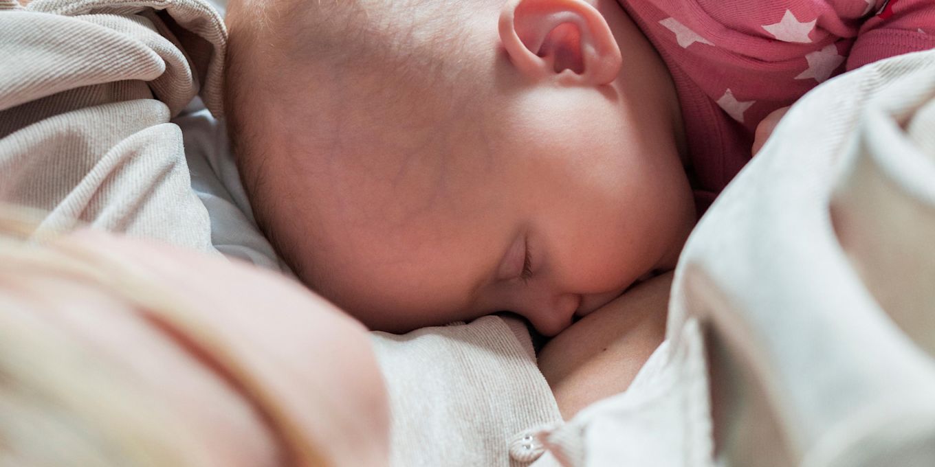 Spädbarn i rosa sparkdräkt ligger i sängen och ammar vid sin mammas bröst.
