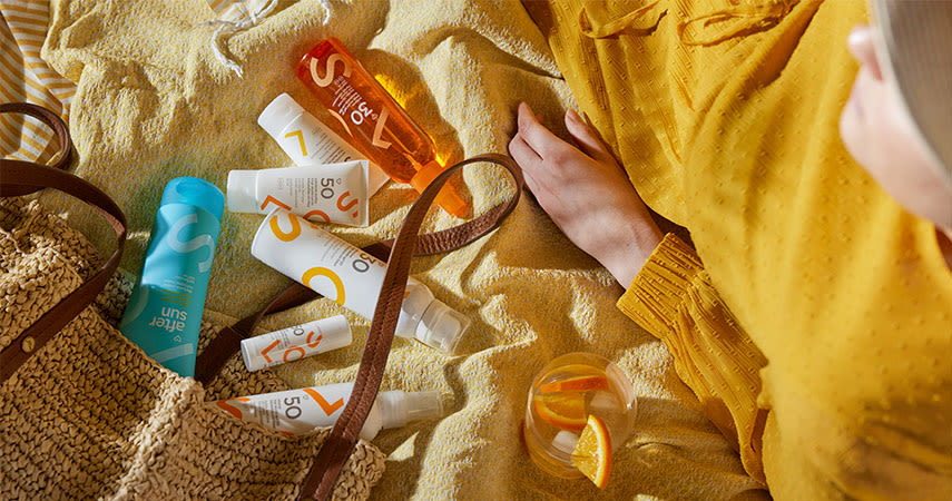 En kvinna i gul klänning ligger på ett badlakan. Ur hennes väska kommer flera produkter från Apotek Hjärtats sortiment med produkter för solskydd.