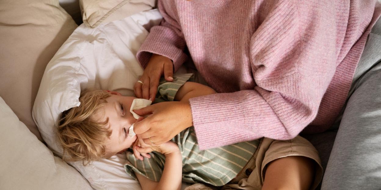 Barn ligger i en säng medan en vuxen torkar näsan med en näsduk. Bilden illustrerar vård av sjuka barn i hemmet och de utmaningar som många familjer möter under vabruari.