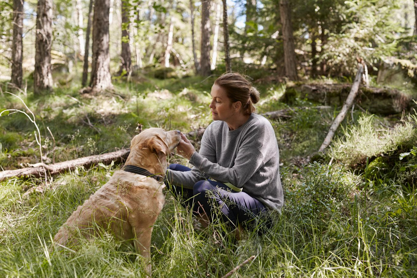 Kvinna sitter i högt gräs ute i skogen tillsammans med en hund som hon klappar på nosen.