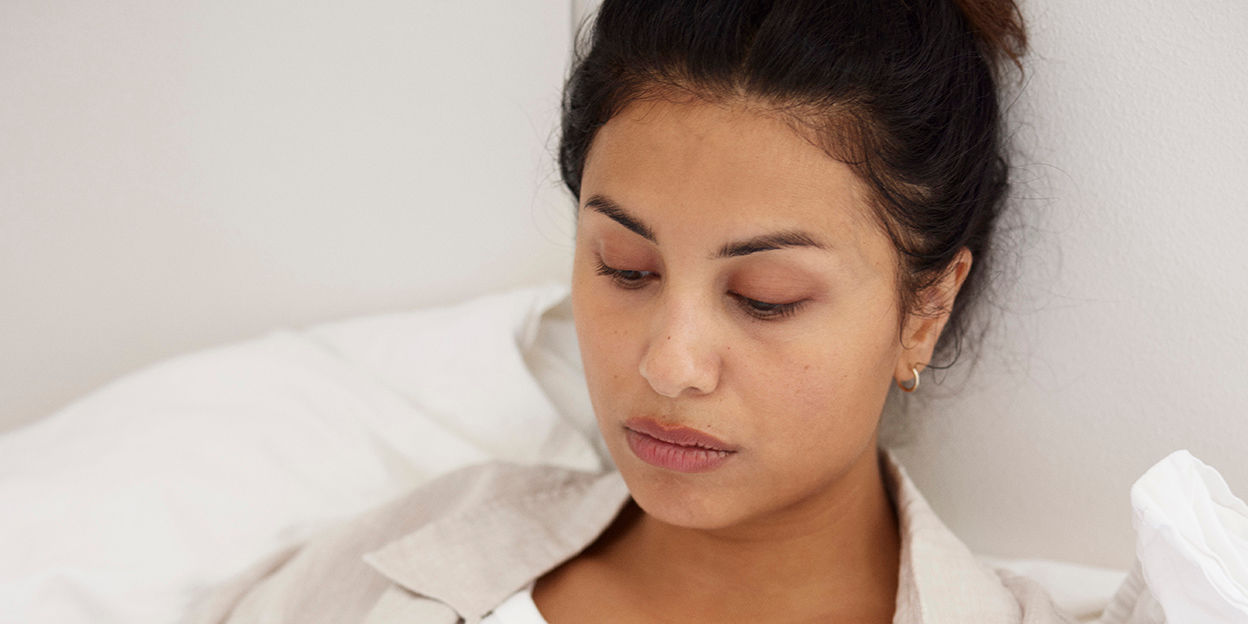 Kvinna sitter i sängen och kollar snett nedåt. 