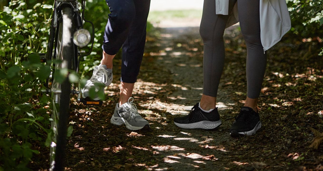 Närbild på två personers smalben och fötter när de står på en skogsstig i träningsskor och träningsbyxor, ena personen har en cykel med sig.