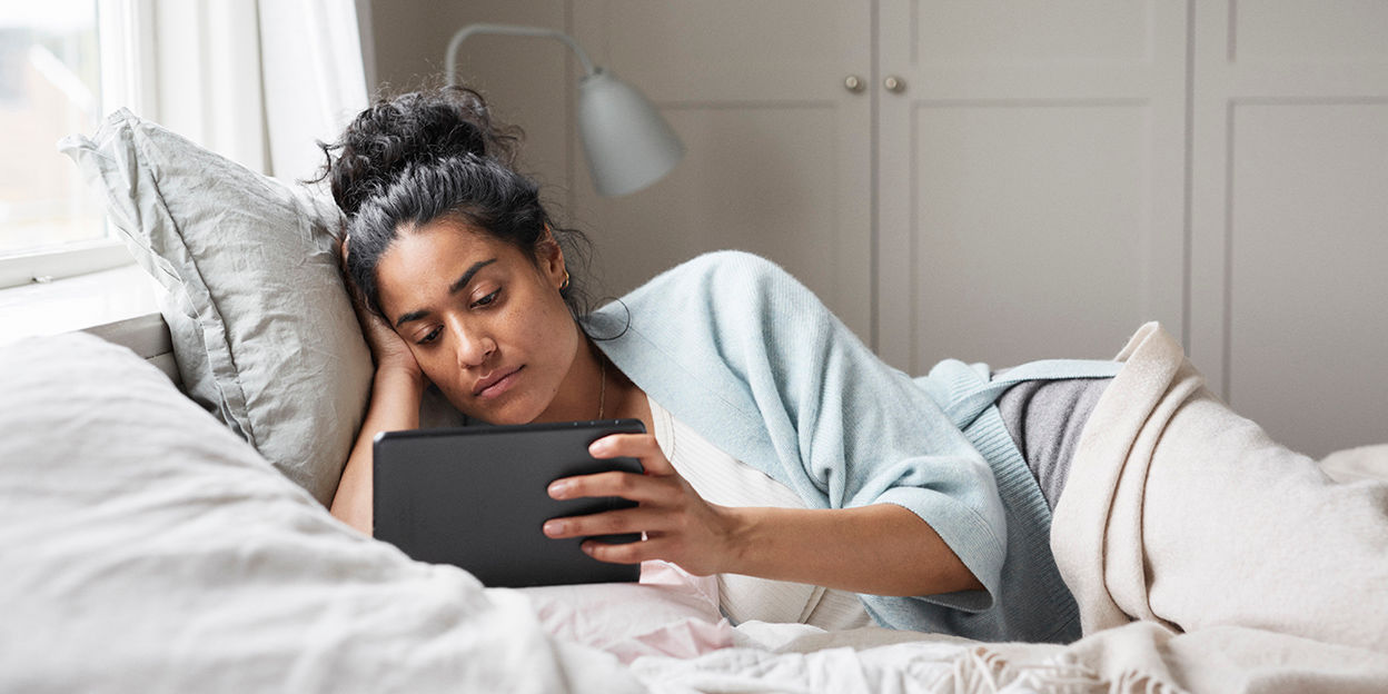 Kvinna ligger i sängen och vilar. Hon tittar på en surfplatta som hon håller med ena handen.