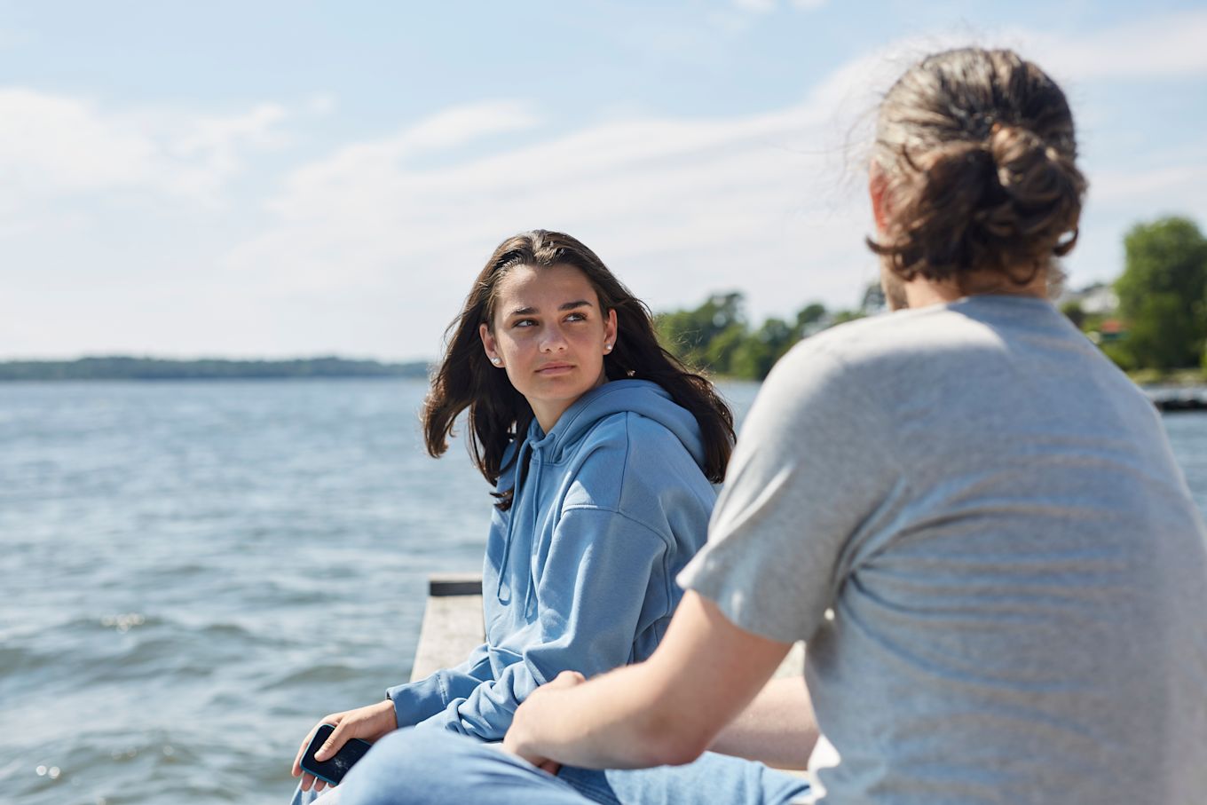 Tonårstjej sitter med pappa utomhus på sommaren på en brygga vid vattnet och pratar.