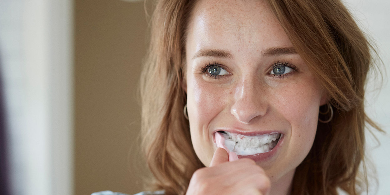 Kvinna borstar tänder och ler. Hon kollar snett uppåt.