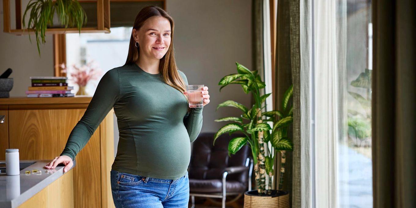 Person står i ett kök och håller ett glas vatten med kosttillskott medan dagsljus strömmar in genom fönstret.