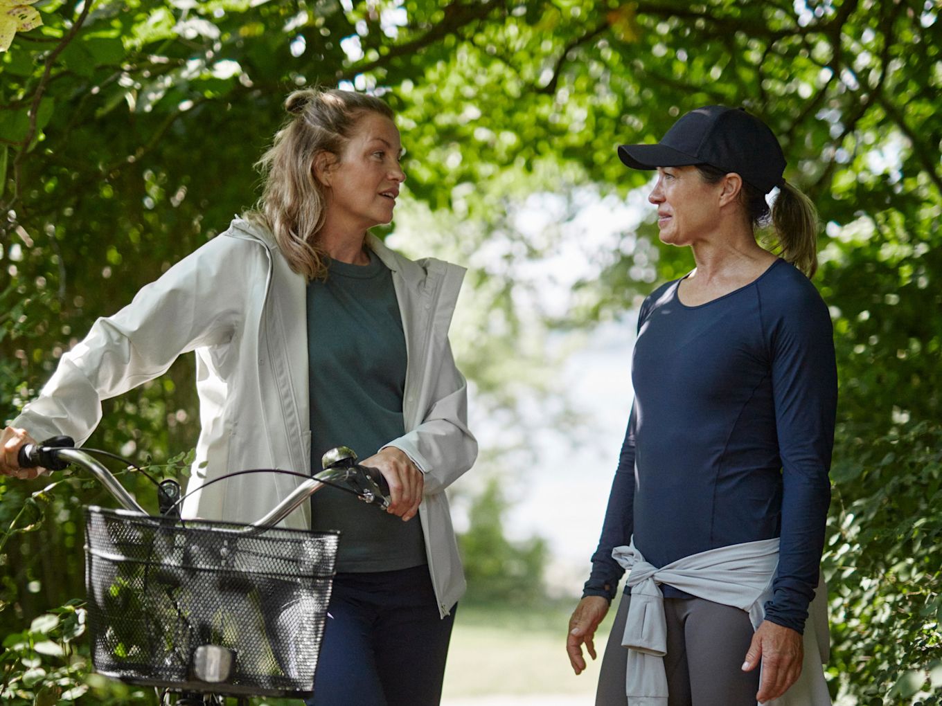Två kvinnor står utomhus i skogen och pratar, iklädda träningskläder. Den ena kvinnan håller i en cykel.