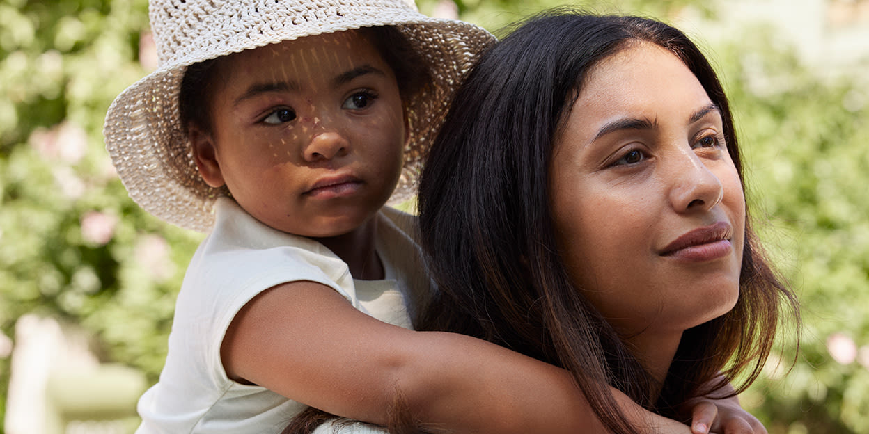 Mamma har sin dotter på ryggen. De är utomhus i solen.