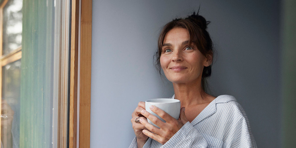 Kvinna står vid ett fönster, iklädd pyjamas med en temugg i händerna. Hon tittar åt sidan och ser nöjd ut.