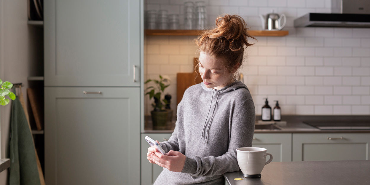 Tjej står i köket och kollar ned på sin telefon. Hon har en kopp te bredvid sig. 