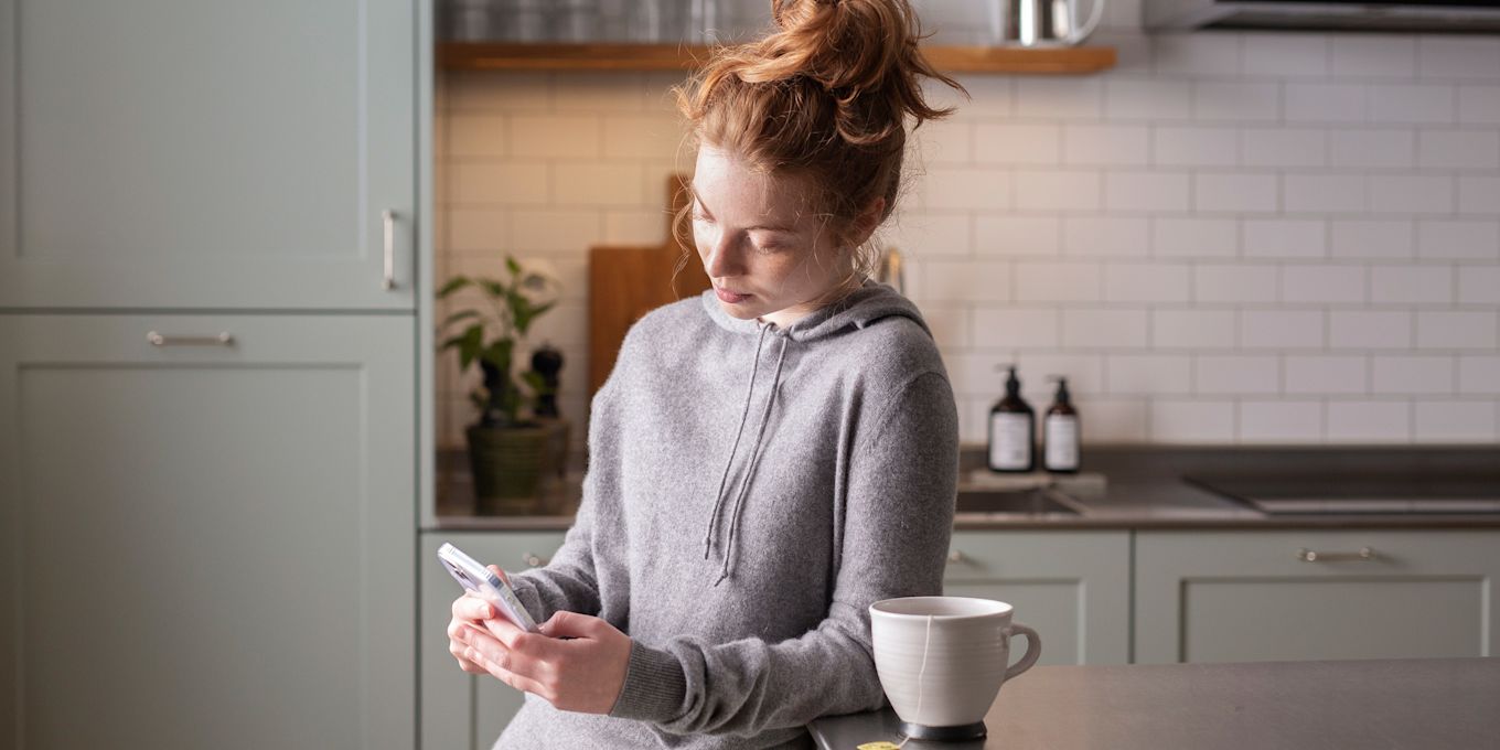 Tjej står i köket och lutar sig mot köksbänken samtidigt som hon tittar på sin telefon. Det står en kopp te bredvid.