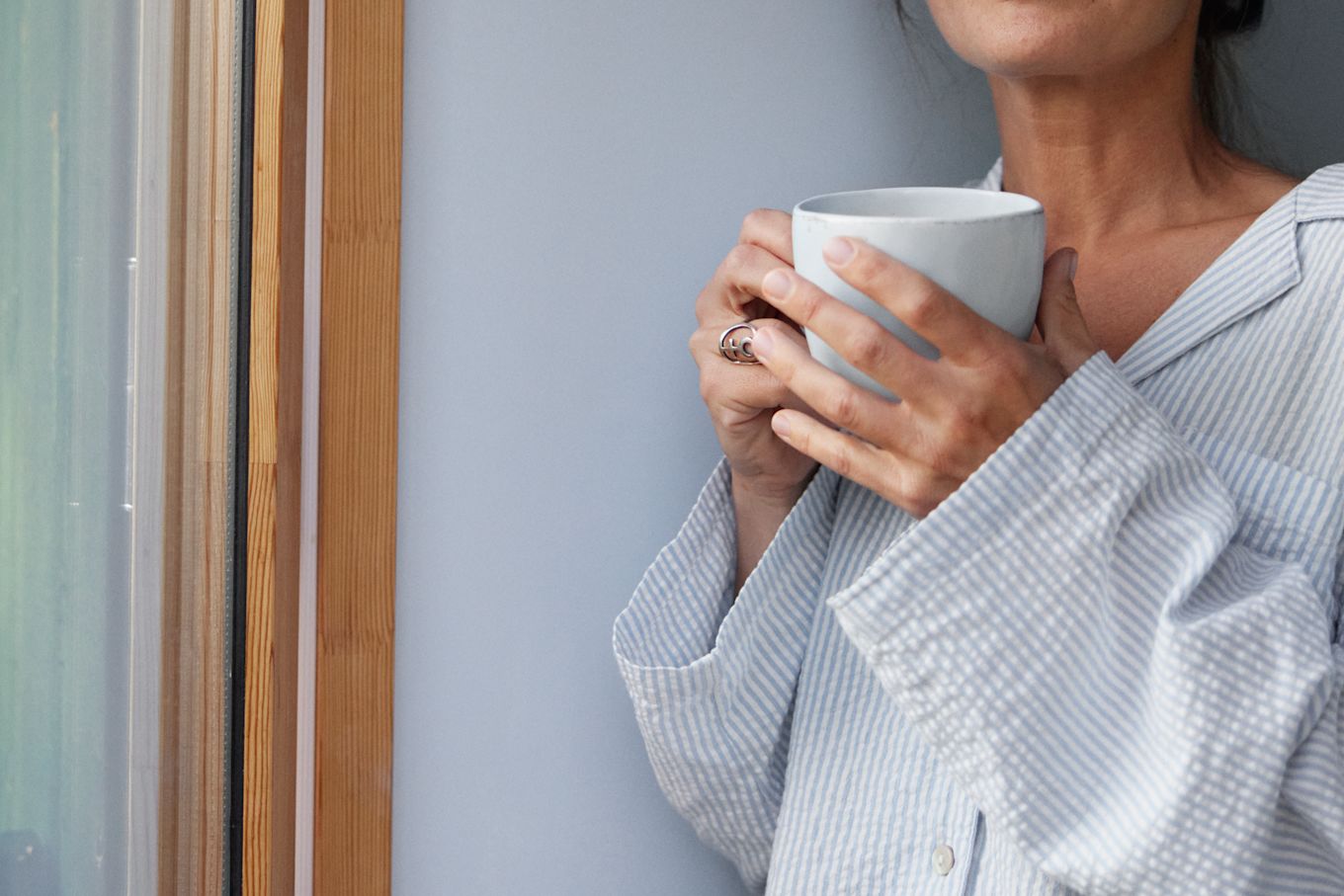 Kvinna iklädd blårandig pyjamas står lutad mot en vägg intill ett fönster, hon håller i en vit mugg.