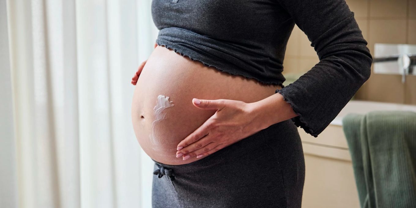 Person smörjer in den gravida magen med hudkräm i ett badrum. Bilden illustrerar hudvård under graviditeten och hur huden kan tas om hand när magen växer.