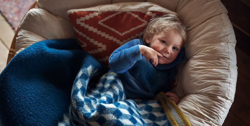 Liten pojke ligger och myser med filtar i en fotölj. 