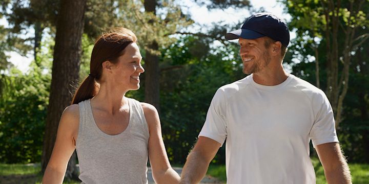 Kvinna  och man går en promenad en solig dag genom en park. De håller varandra i handen och tittar på varandra och ler