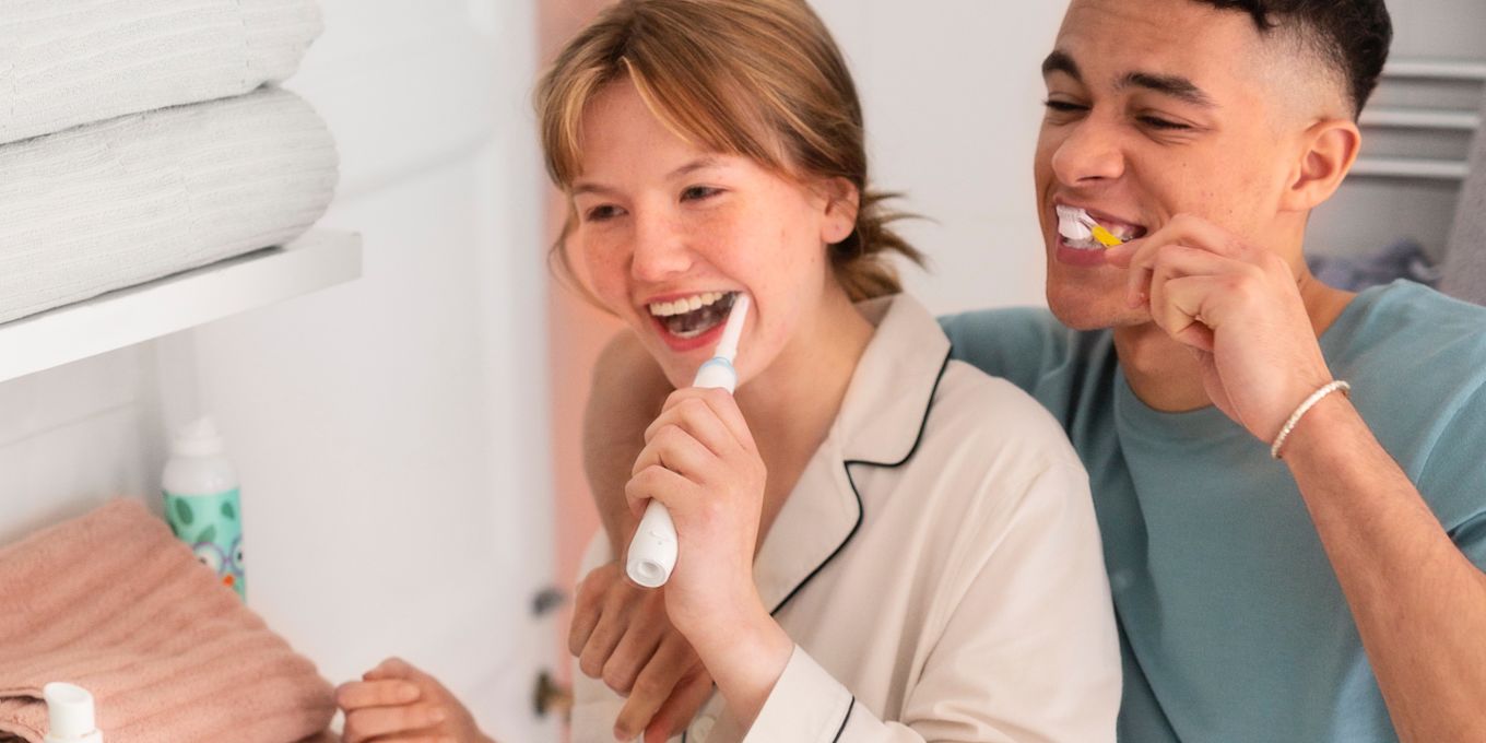 En ung tjej och kille står i badrummet och borstar tänderna tillsammans och skrattar. Tjejen använder en eltandborste medan killen använder en vanlig manuell tandborste.