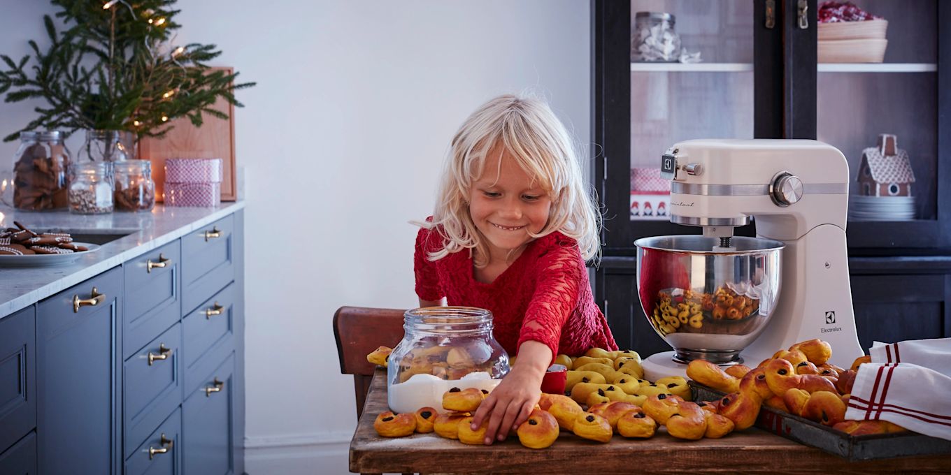 Ett barn sträcker sig efter nybakade lussekatter på ett köksbord med en köksmaskin.