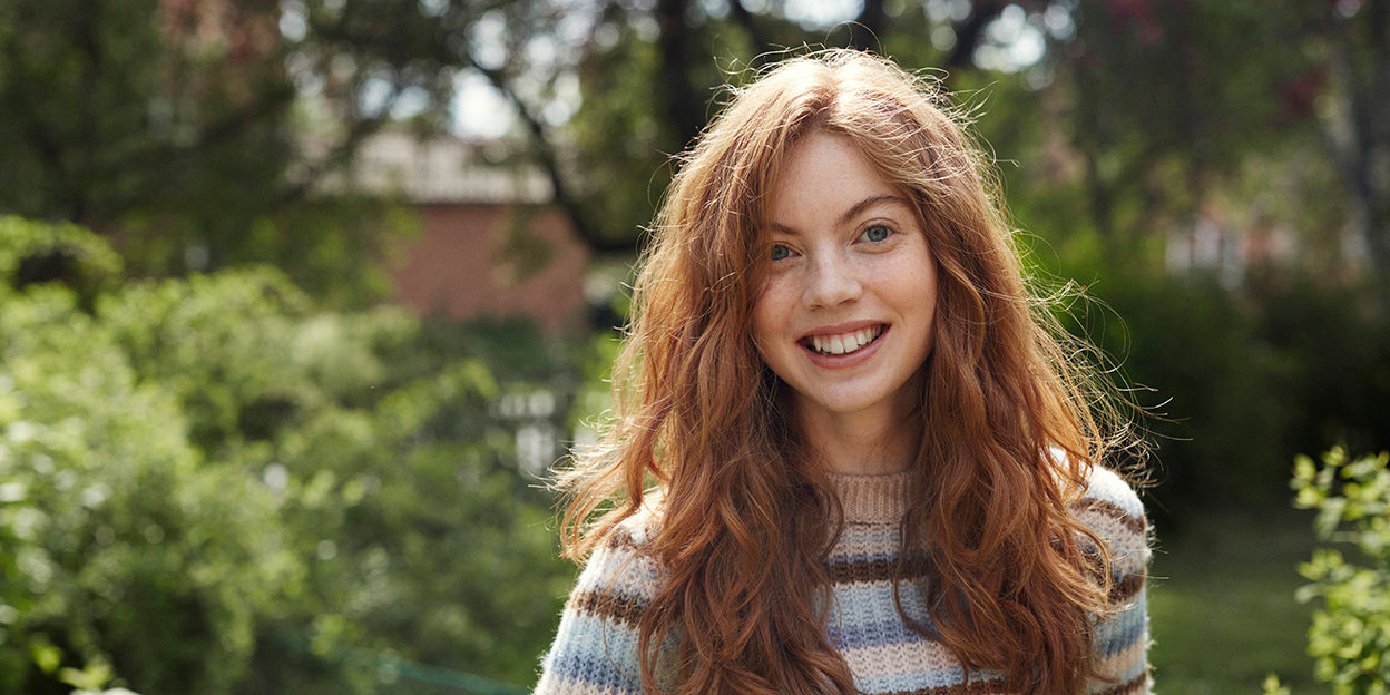 En glad ung kvinna vistas ute i trädgårdsmiljö en solig dag.