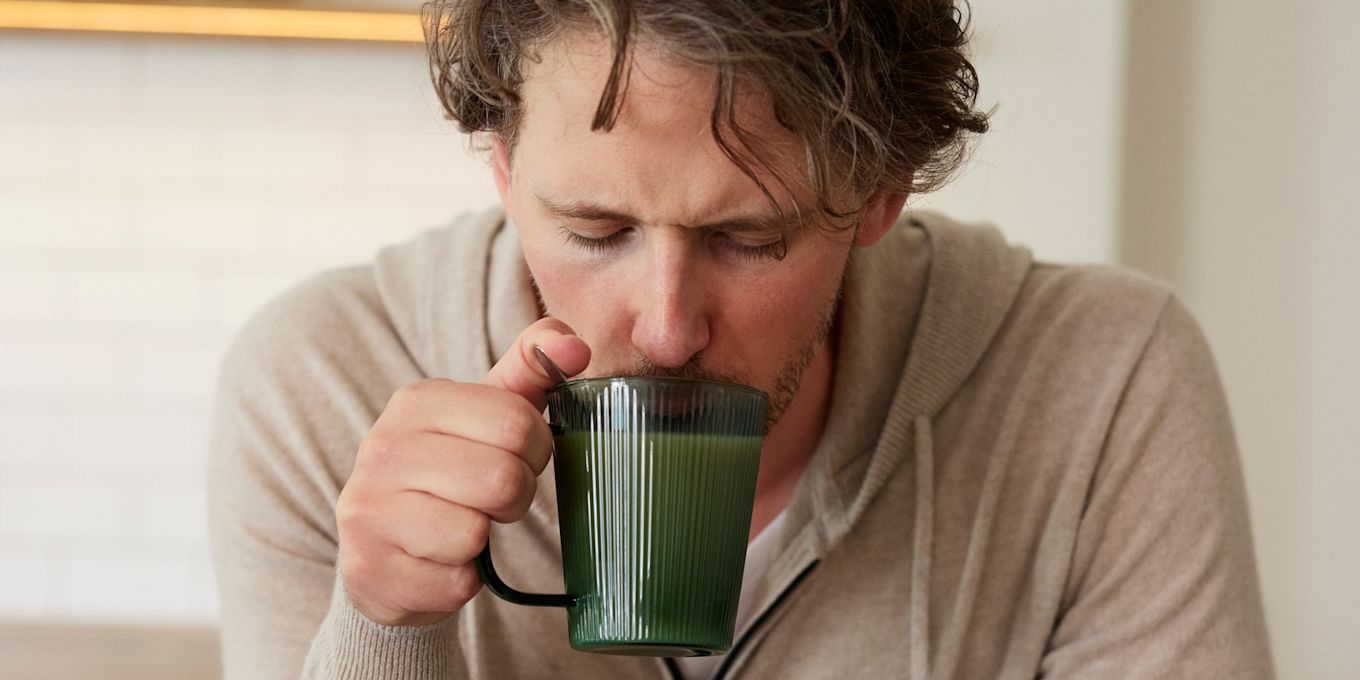 Kille dricker varm dryck ur en grön glasmugg. Han tittar ner i muggen och ser lite trött ut.