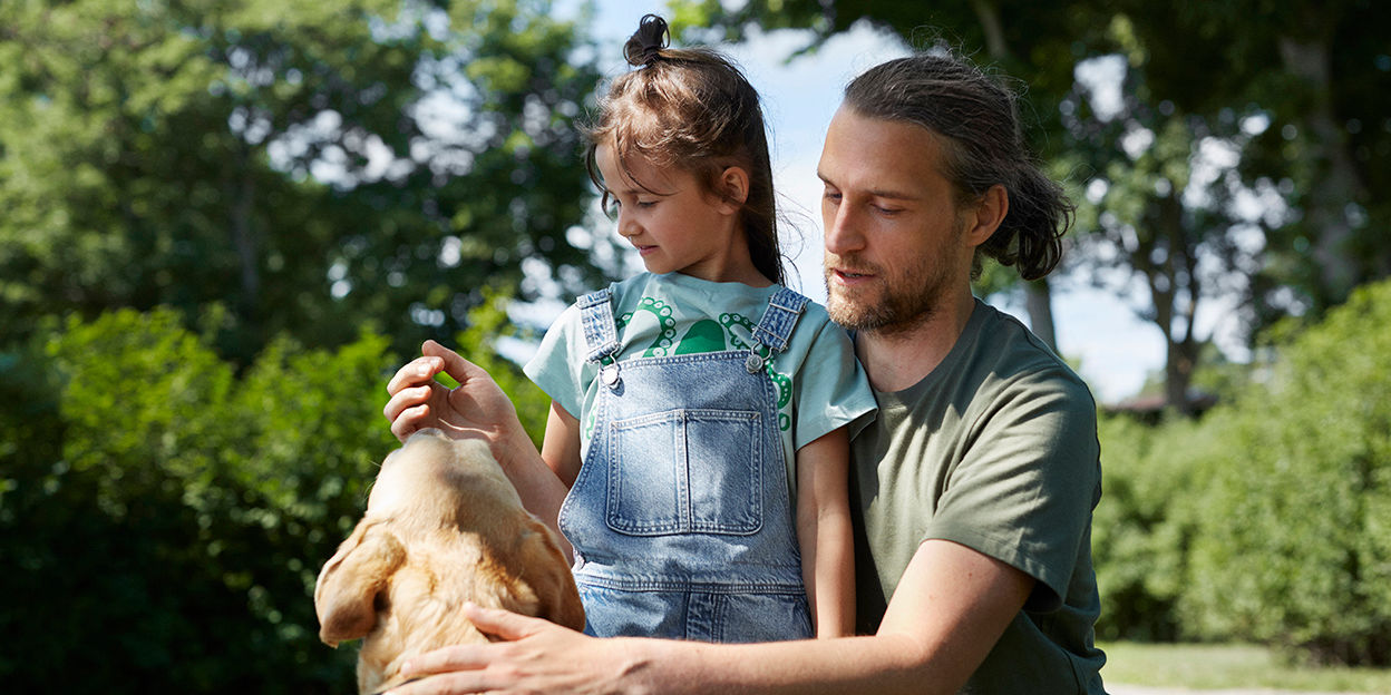 Pappa, dotter och hund är ute i parken en sommardag.