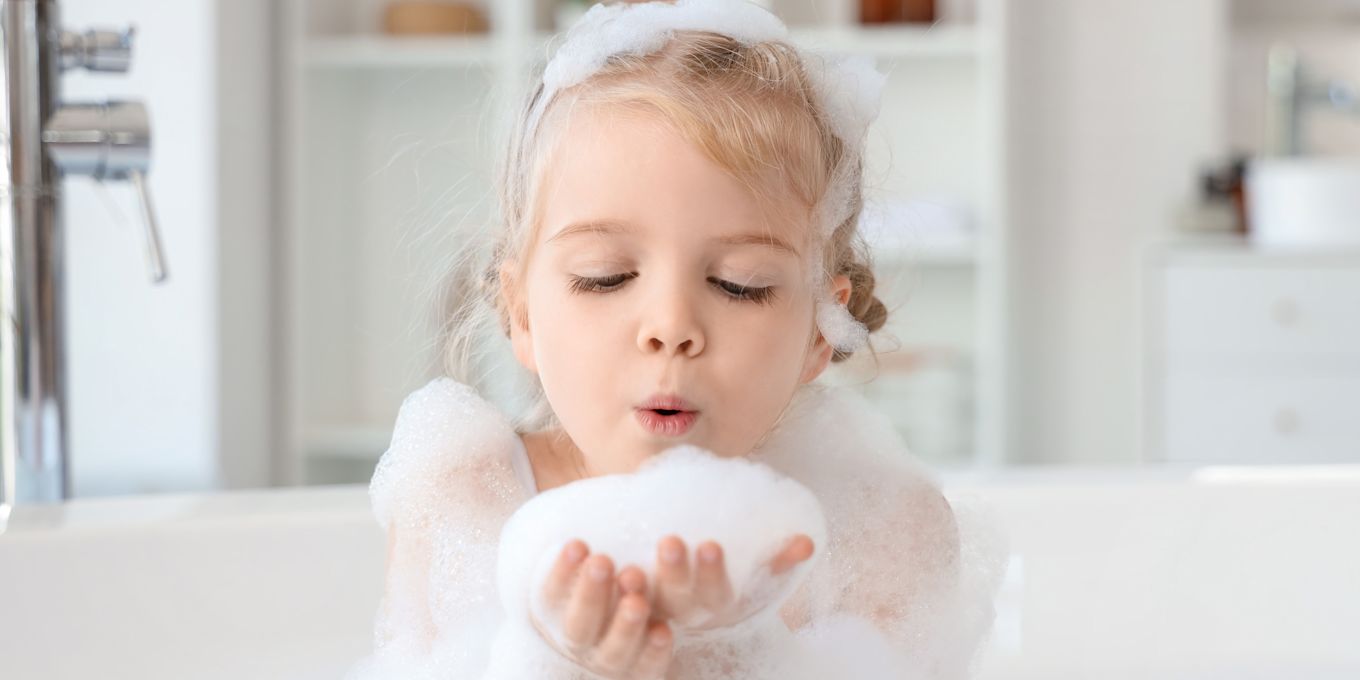 En flicka sitter i badkaret och håller badskum i handen som hon blåser på.