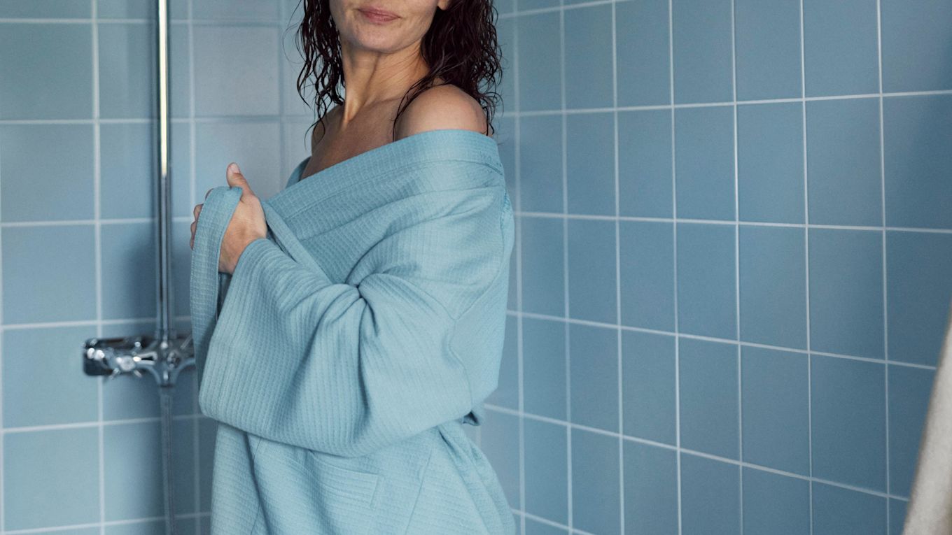 Kvinna står i ett blått badrum, nyduschad och sveper in sig i en morgonrock.