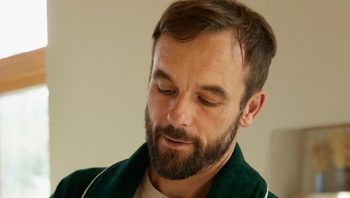 Man med brunt hår och skägg står och tittar ner i marken.