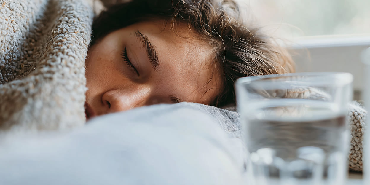 Kvinna med feber sover under en filt med ett glas vatten framför sig.