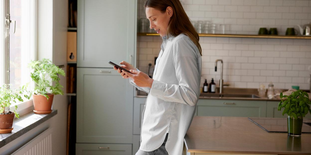 Kvinna står i köket och tittar ner på sin mobiltelefon.