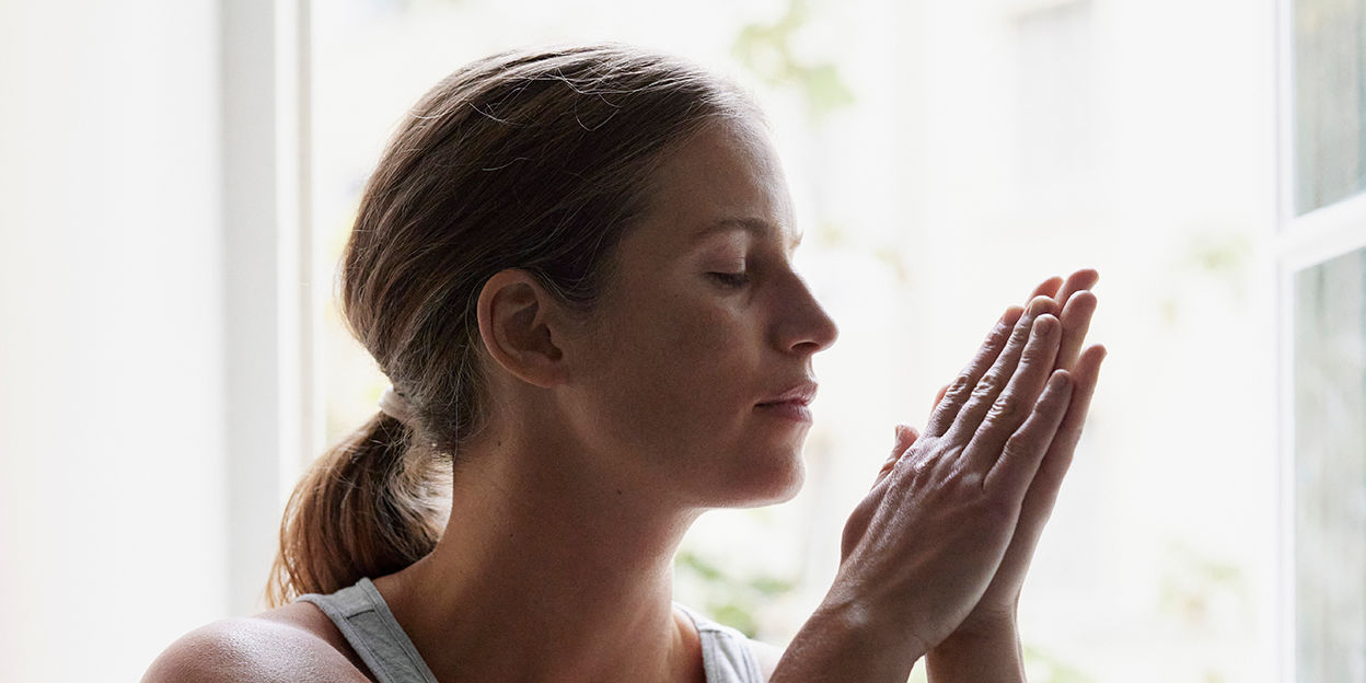 Kvinna står vid fönster. Står i profil, håller händerna ihop och kollar snett nedåt.