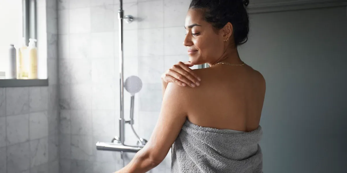 Kvinna står i badrum, invirad i handduk och smörjer in sin axel med lotion.