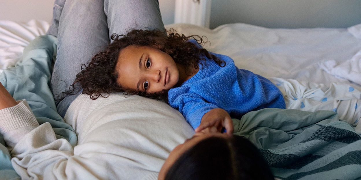 Mamma och dotter ligger i sängen. Dottern vilar sitt huvud mot mamman.