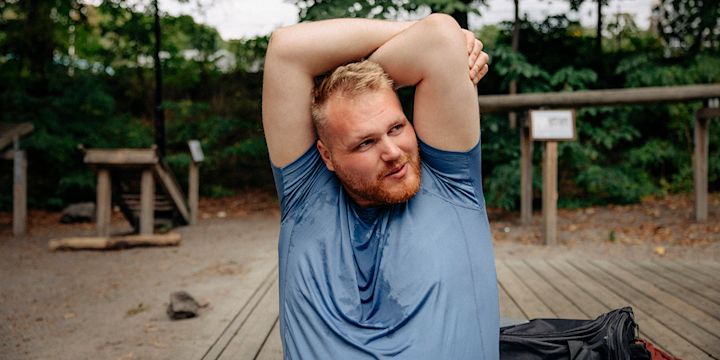 Kille sitter på ett utegym och håller upp armarna för att stretcha triceps. Han har en blå t-shirt som är lite svettig.