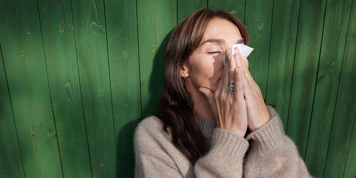 Kvinna sitter utomhus, lutad mot en grön vägg och snyter sig.