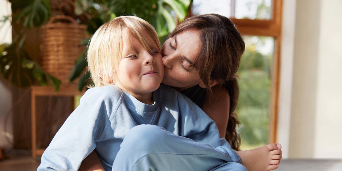 Mamma och son sitter tillsammans på golvet och leker och myser, mamma pussar sonen på kinden.