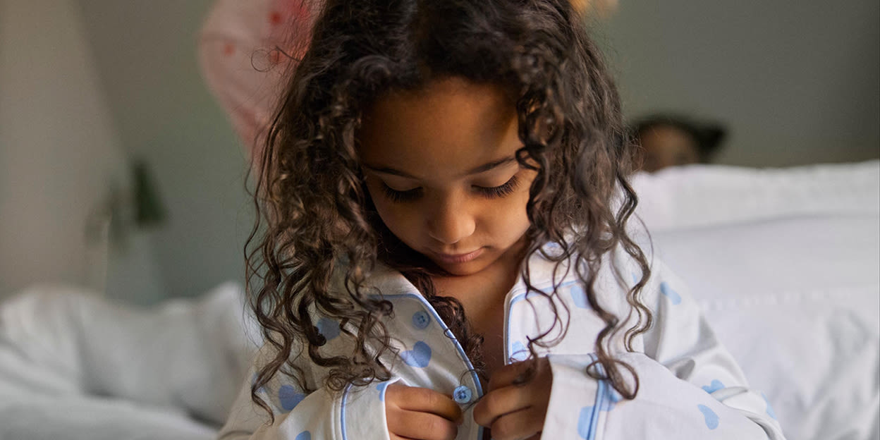 Liten flicka i 5-års åldern med lockigt mörkbrunt hår knäpper sin pyjamas som är vit med blå prickar, hon står i ett sovrum.