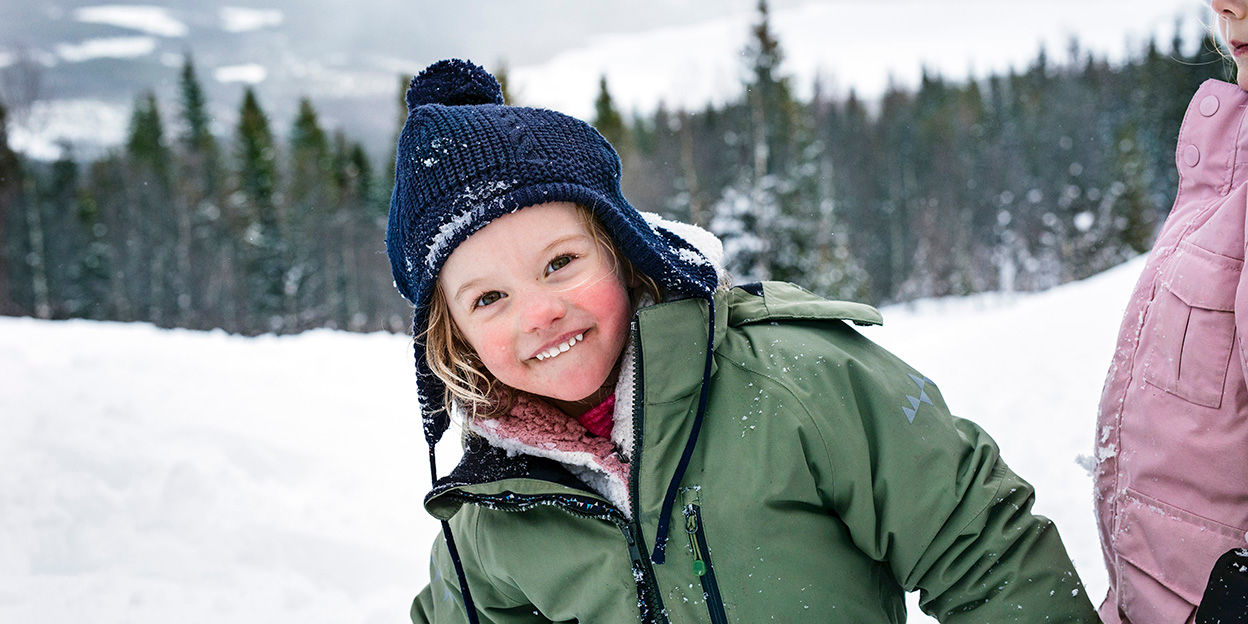 Pojke är ute i snön med vinterkläder på. Hans kinder är röda och han ler stort mot kameran. 