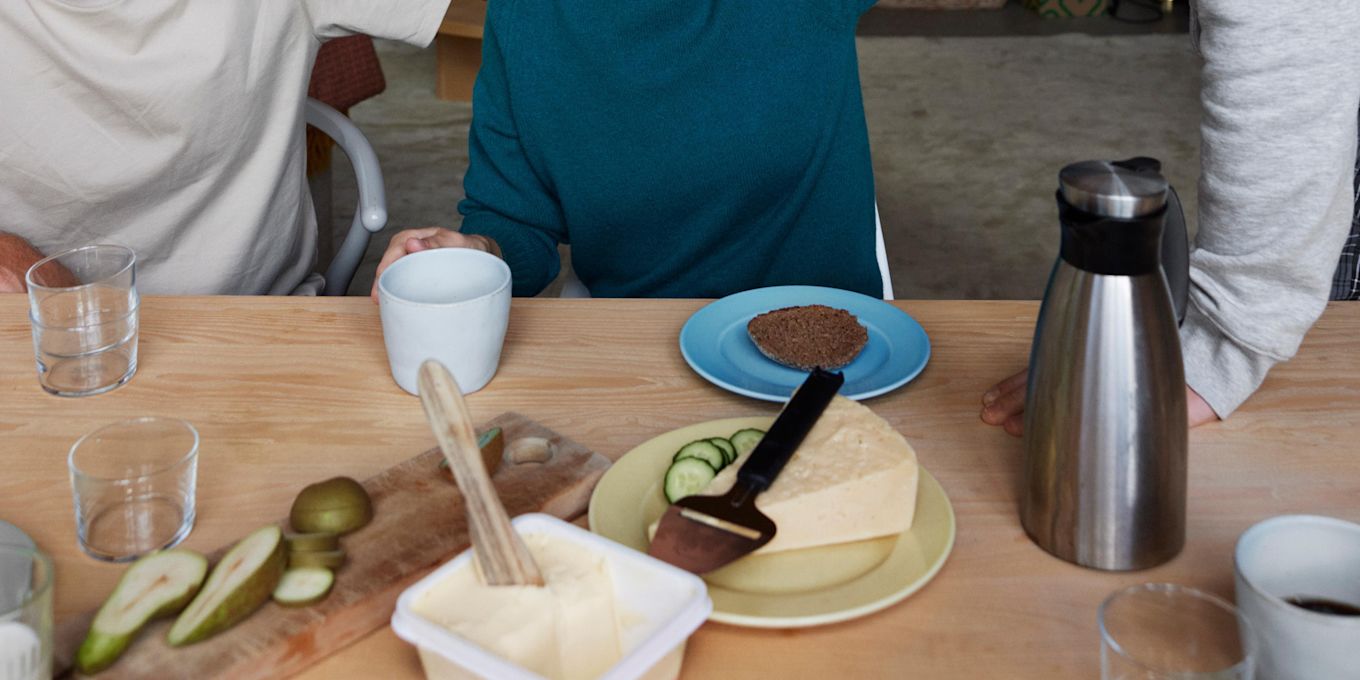 På ett matbord står det ost, smör, kaffekoppar, termos, bröd och glas.
