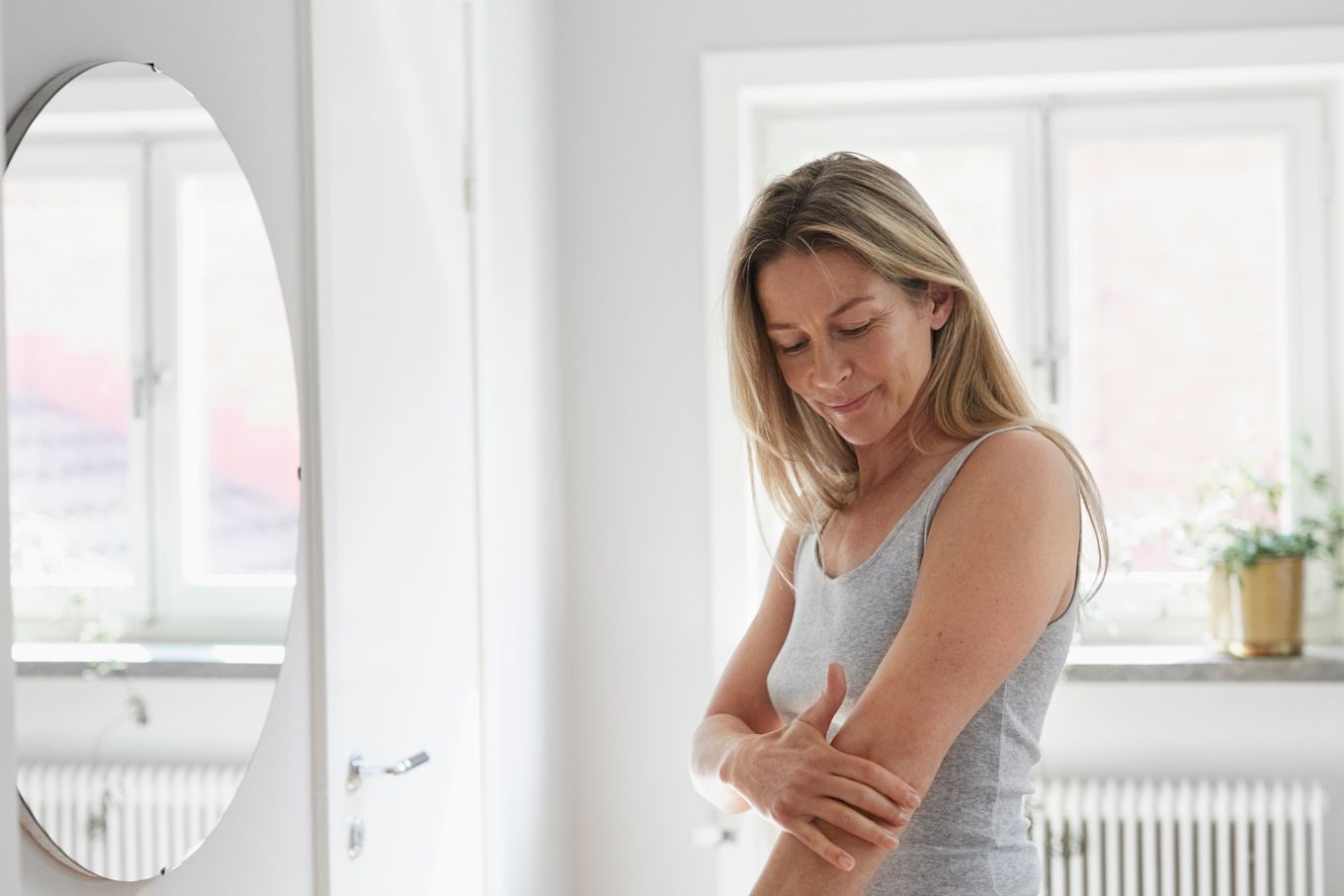 En medelålderskvinna står och smörjer in sin arm framför en spegel, solljuset lyser in i bakgrunden genom fönstret.